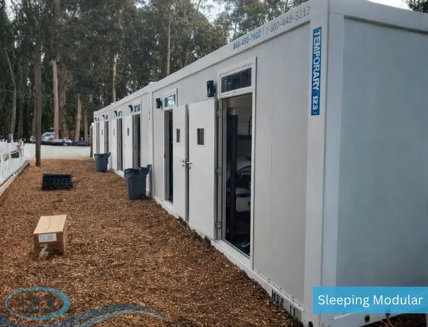 Row of white modular units with open doors on a wood-chip path, set amid tall trees. They convey a sense of practicality and temporary housing.